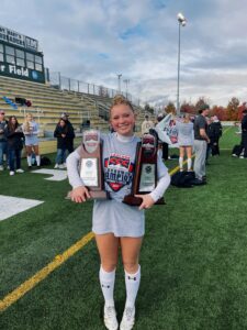 Gracie stands on a soccer field holding two trophies and smiling at the camera.